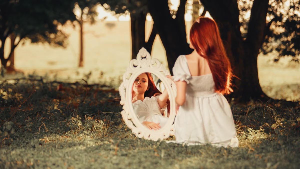 A redhead woman sits outdoors with a mirror reflection, surrounded by nature.